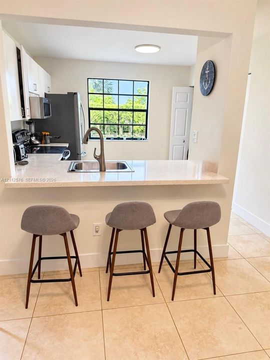 Kitchen featuring light-toned cabinetry, solid surface breakfast bar, stainless steel undermount sink with gooseneck faucet, window with black grid framing, and neutral tile flooring