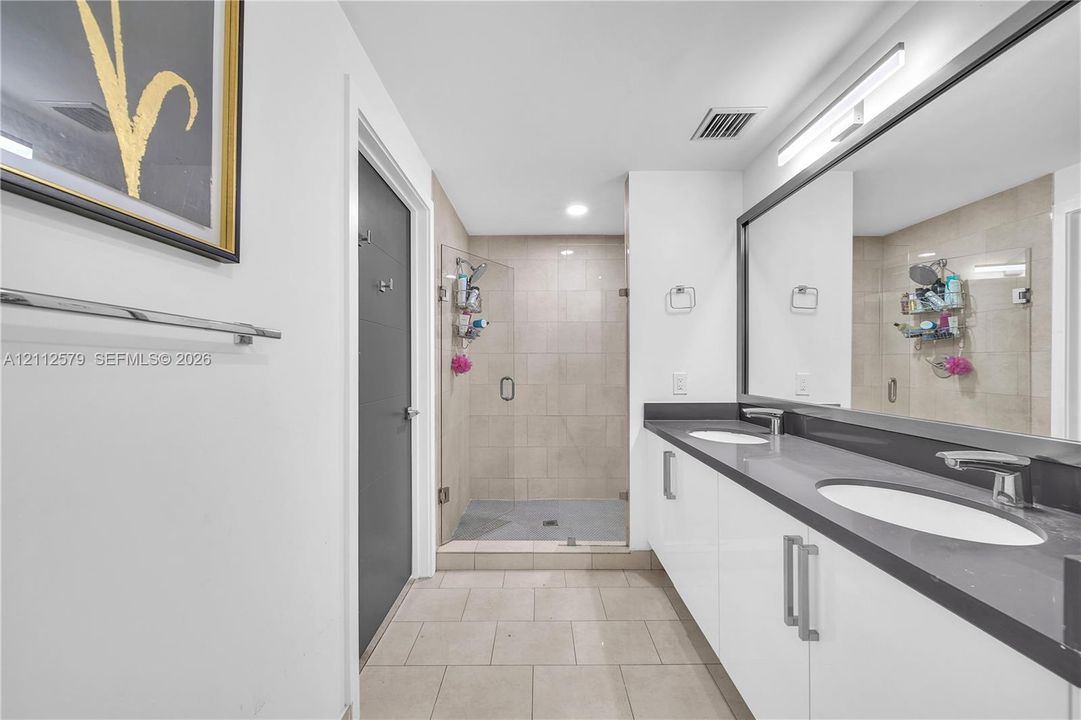 Dual-sink vanity with high-gloss cabinetry and stone-finish countertop, frameless glass shower enclosure with neutral-toned wall tile, and full-width vanity mirror with integrated lighting