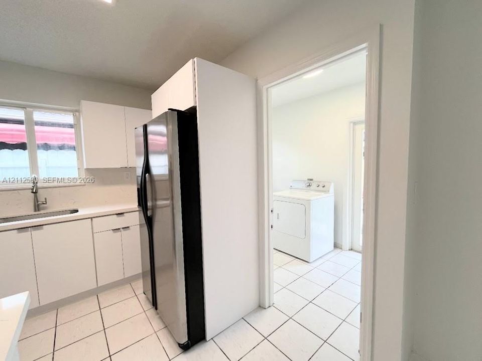 Kitchen featuring white cabinetry, light-toned countertops, a stainless steel refrigerator, and tiled flooring extending into an additional room containing laundry hookups