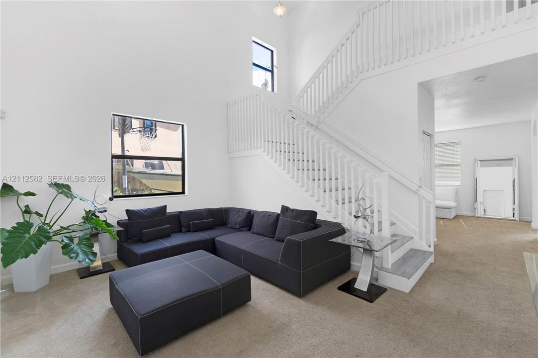 High-ceiling living area featuring a central staircase with white wooden balusters, large windows, and carpeted flooring