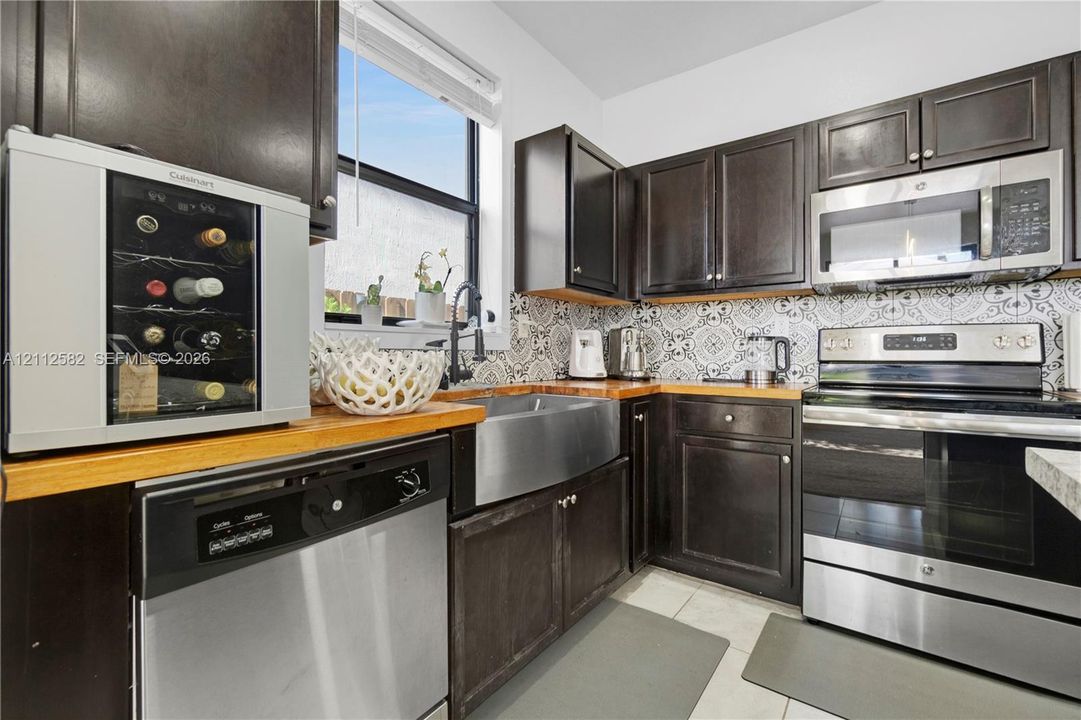 Kitchen featuring dark-finish cabinetry, butcher block countertops, patterned tile backsplash, stainless steel farmhouse-style sink, and stainless steel appliances