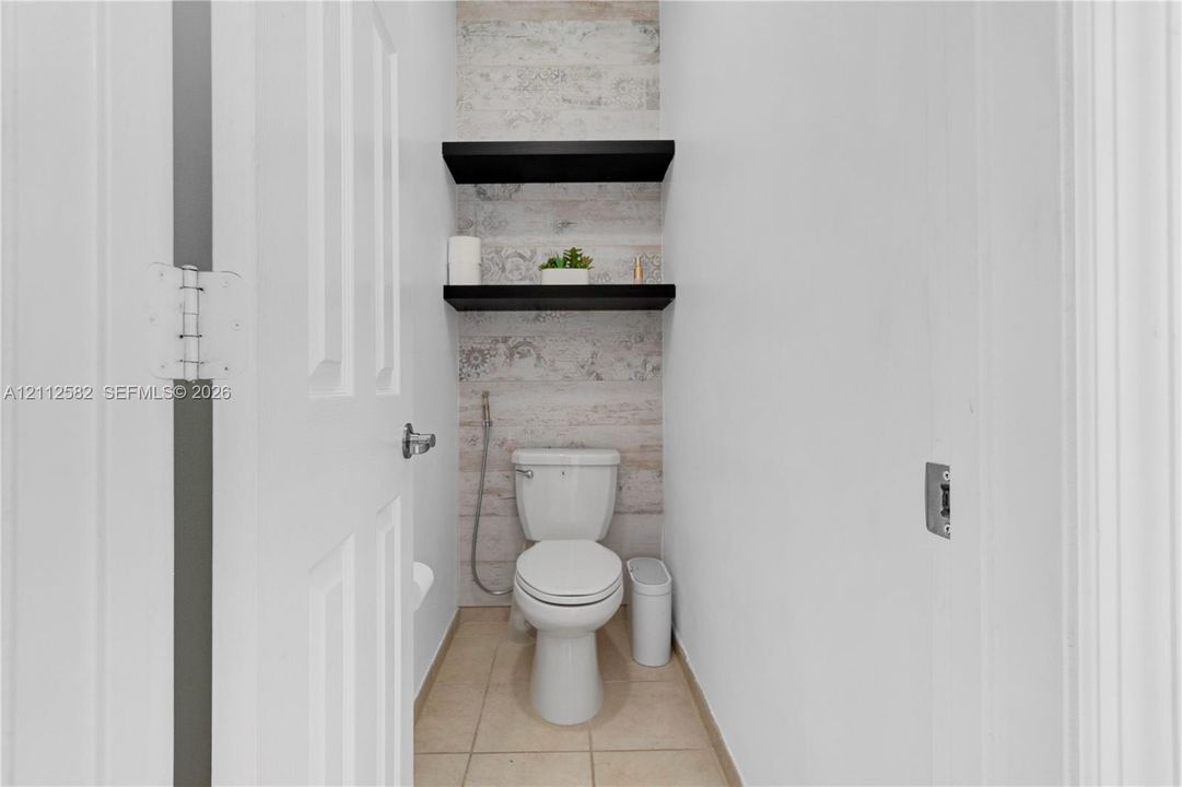 Restroom featuring ceramic tile flooring, two black floating shelves, decorative wall surfacing, and a wall-mounted bidet sprayer