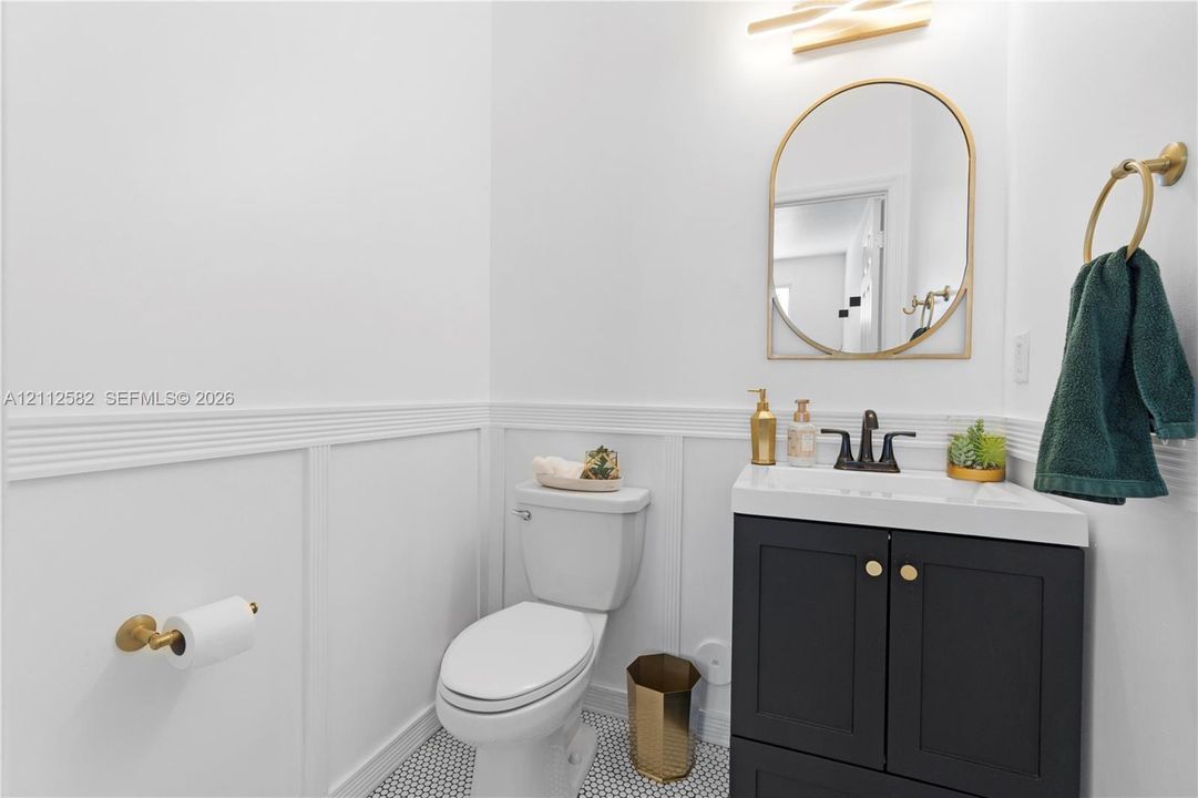 Powder room featuring wainscoting, a dark vanity cabinet with a stone-top sink, arched gold-rimmed mirror, patterned floor tile, and gold hardware fixtures
