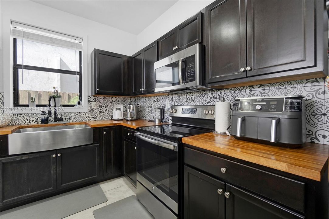 Kitchen featuring dark cabinetry, butcher block countertops, stainless steel farmhouse sink with a gooseneck faucet, stainless steel appliances, and patterned tile backsplash