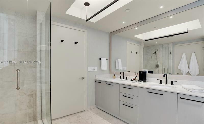 Dual-sink vanity with light-gray cabinetry, stone countertops, matte black fixtures, full-width mirror, and walk-in tiled shower with a glass enclosure