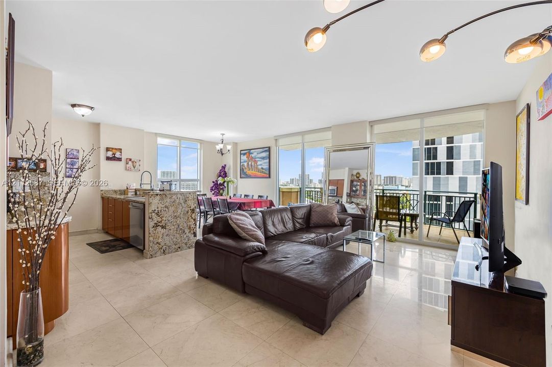 Open-concept interior featuring light-toned tile flooring, floor-to-ceiling glass balcony doors, kitchen island with stone-surface countertop, and multi-arm statement lighting