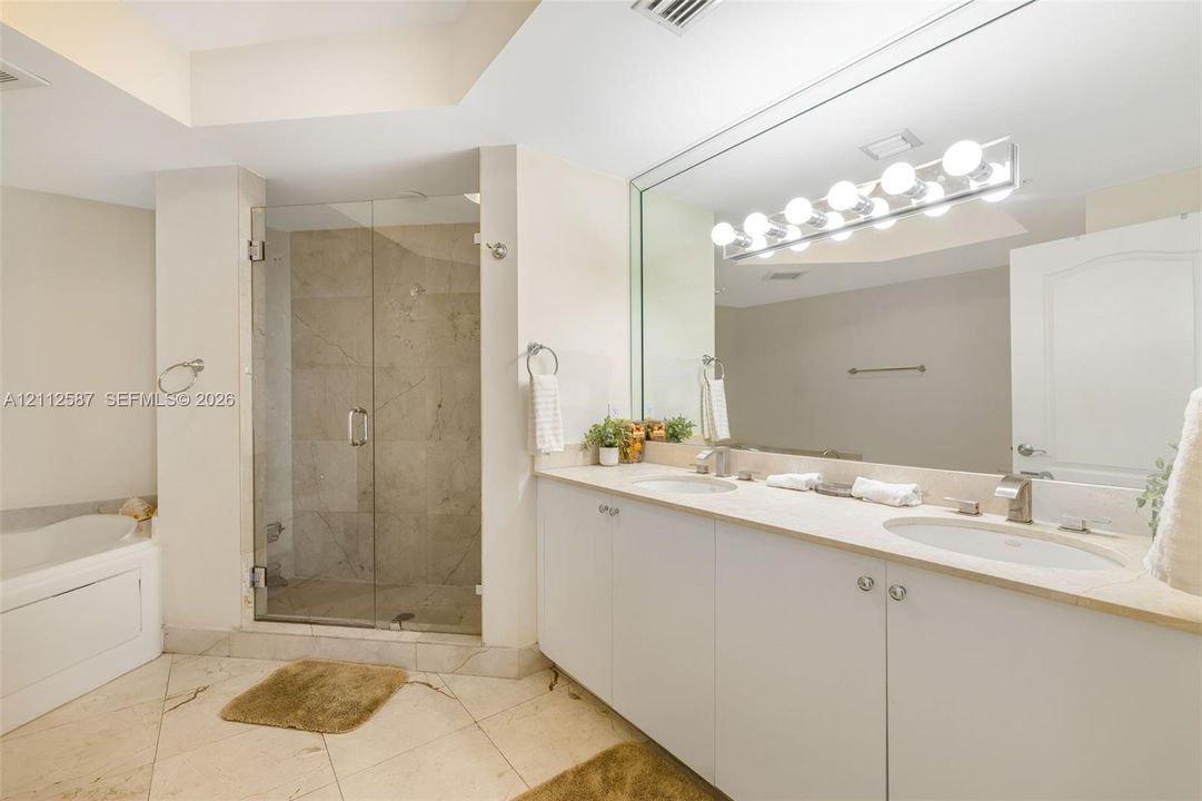 Double vanity with integrated sinks, stone-surface countertop, glass-enclosed shower featuring tile surround, oversized wall mirror, and soaking tub