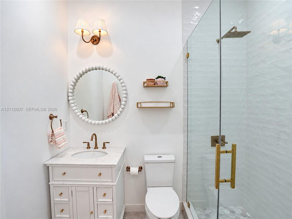White vanity cabinet with a stone countertop, wall-mounted dual-bulb light fixture, circular bead-detail mirror, and a glass-enclosed shower with a rainfall showerhead and polished metal hardware