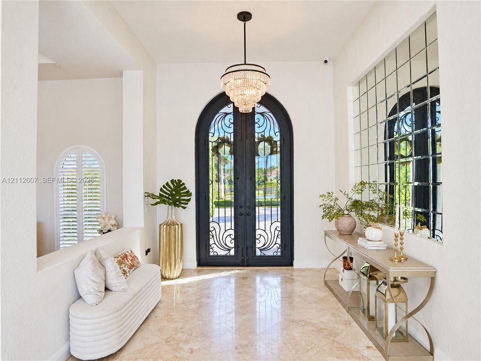 Foyer featuring arched double iron entry doors, light-toned stone flooring, a multi-tiered crystal chandelier, and an expansive grid-pattern wall mirror