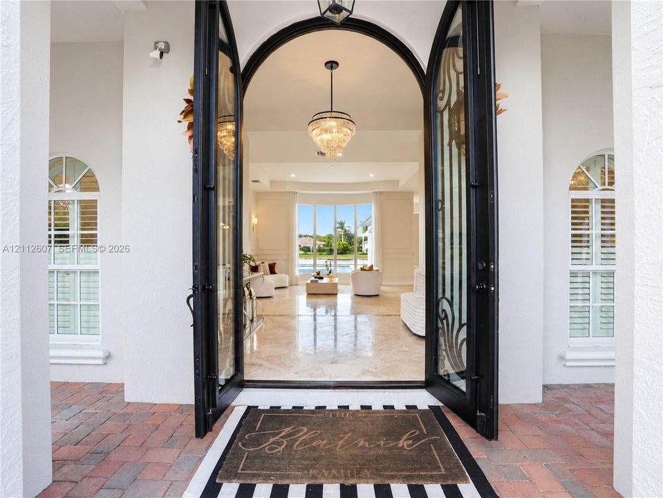Arched double entry doors with decorative iron detailing, flanked by arched windows with plantation shutters, polished stone flooring, and a multi-tiered crystal pendant light fixture