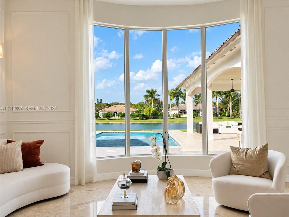 Curved floor-to-ceiling windows, wall paneling, and polished stone flooring, featuring exterior access to a covered lanai, private pool, and water frontage
