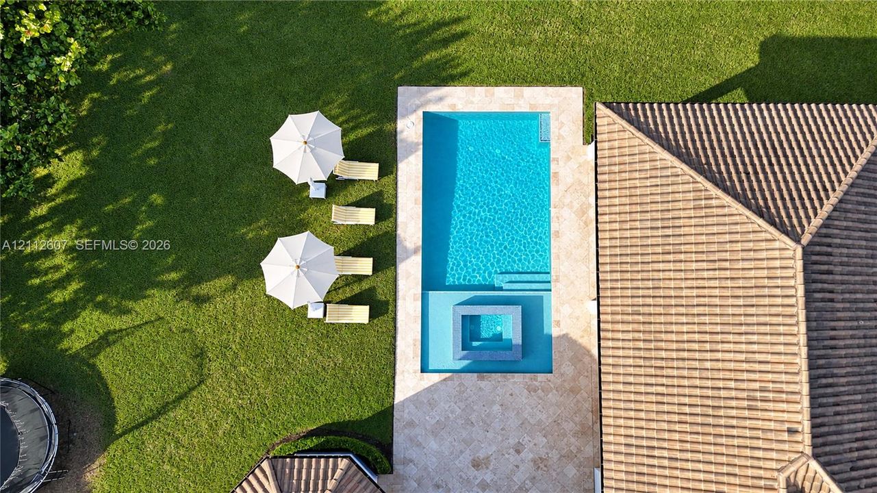 Aerial perspective of a rectangular swimming pool with an integrated spa, featuring stone-finish decking, tile roofing, and a manicured lawn