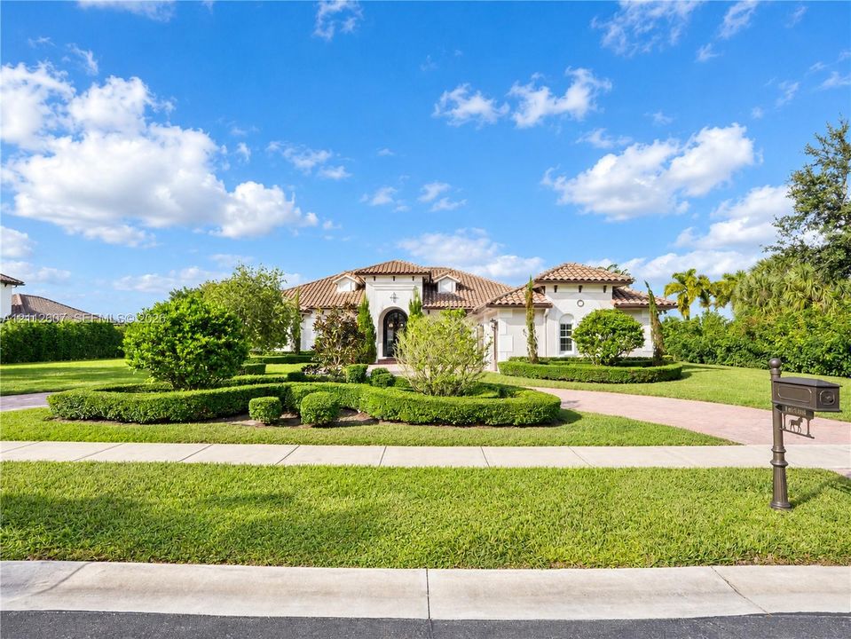 Stucco exterior featuring a clay tile roof, arched entryway, paved circular driveway, and manicured landscaping with shaped hedging