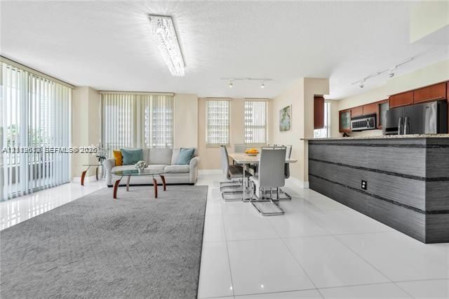 Open-concept interior featuring large-format tile flooring, a kitchen breakfast bar with wood-finish cabinetry, built-in stainless steel appliances, and floor-to-ceiling windows with vertical blinds