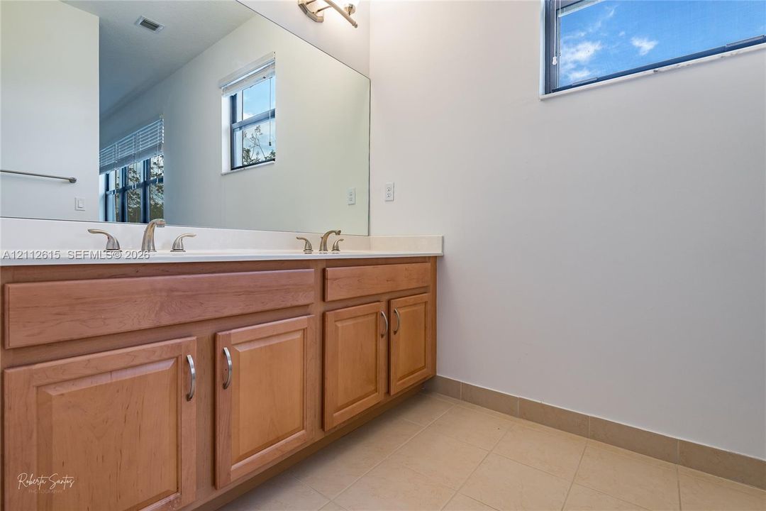 Double-sink wood vanity featuring recessed-panel cabinetry, light-toned countertop, wall-to-wall mirror, and tile flooring