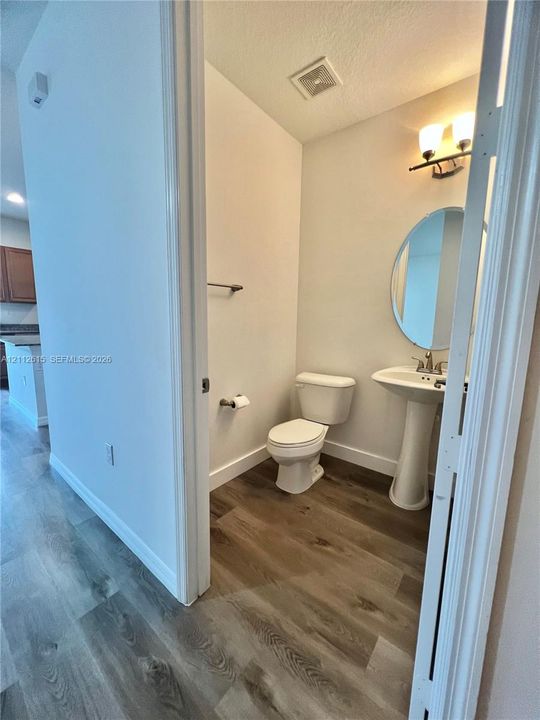 Powder room featuring wood-finish flooring, a white pedestal sink, a circular wall-mounted mirror, and a two-light vanity fixture