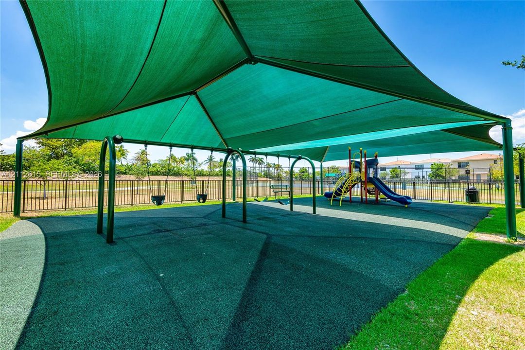 Outdoor recreation area featuring a large overhead sun shade, rubberized ground surfacing, swing sets, and a playground structure enclosed by metal fencing