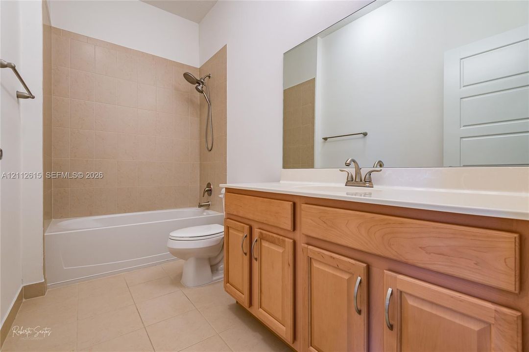 Bathroom featuring a wood-finish vanity with a light-toned countertop, large wall-mounted mirror, ceramic tile flooring, and a tub-shower combination with tiled surround