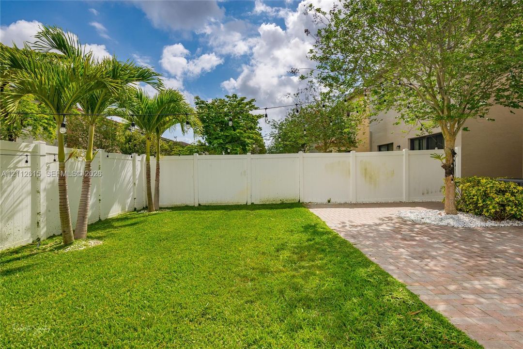 Fenced backyard featuring a manicured lawn, paver patio, mature trees, and palms