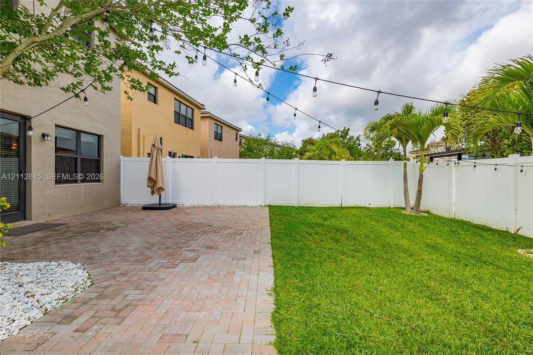 Private outdoor space featuring a paved brick patio, manicured lawn, white vinyl fencing, and overhead string lighting