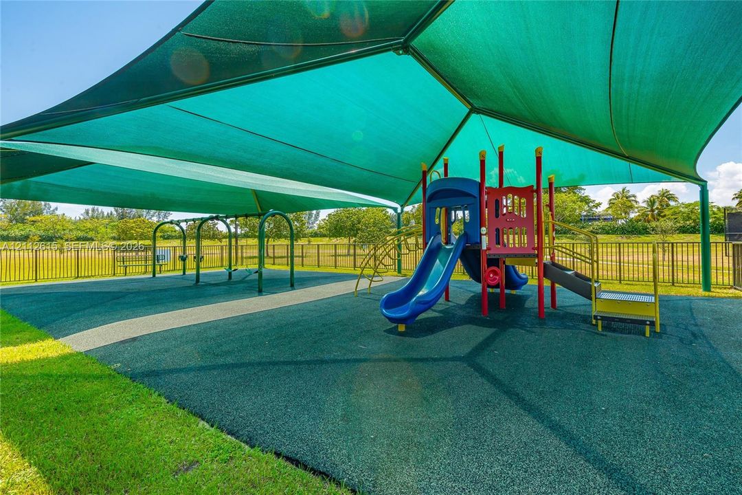 Recreational area featuring a large overhead sun shade, soft-surface ground cover, metal perimeter fencing, multi-station play structure with slides, and swing set