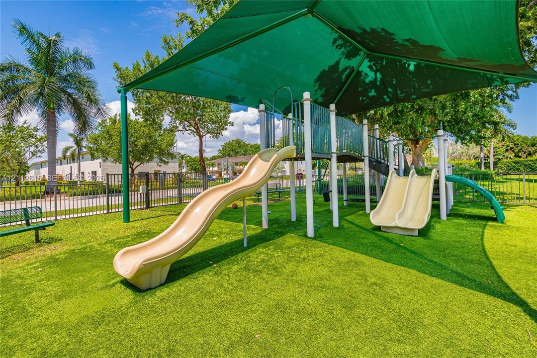 Outdoor recreation area featuring a multi-slide playground structure protected by a large green shade canopy, situated on synthetic turf surfacing and enclosed by perimeter metal fencing