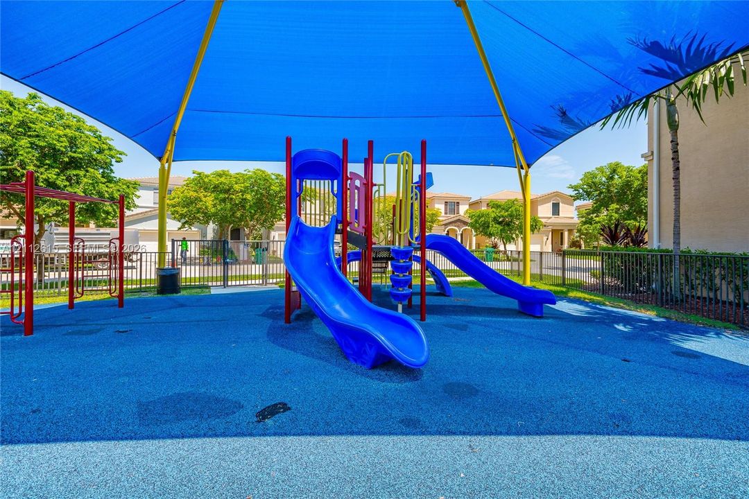 Outdoor recreational area featuring a multi-structure play system, blue poured-rubber flooring, and a large overhead shade canopy