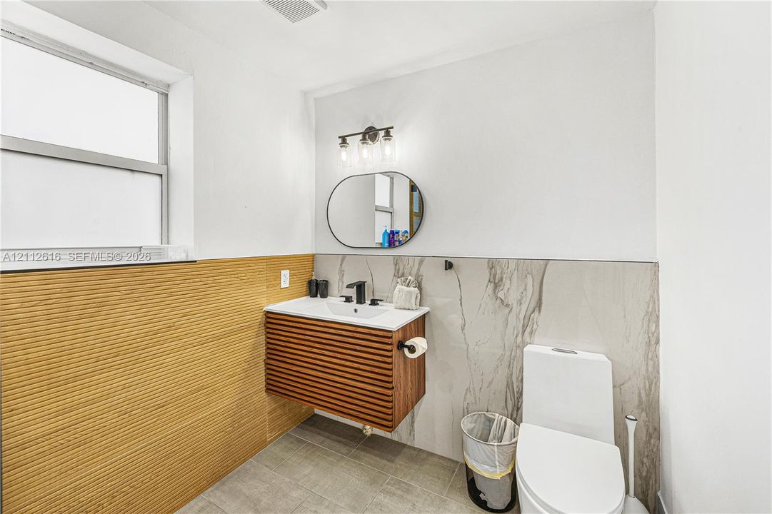 Wall-mounted vanity featuring slatted wood cabinetry, rectangular sink, and matte black faucet, accented by textured wood-finish wall panels, large-format stone-patterned tile wall cladding, and an oval mirror with three-light fixture