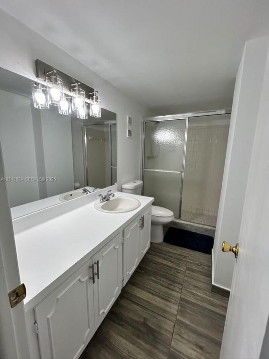 Bathroom featuring a wood-finish tile floor, extended white vanity with a solid-surface countertop, full-width mirror, multi-bulb wall-mounted lighting, and a glass-enclosed shower