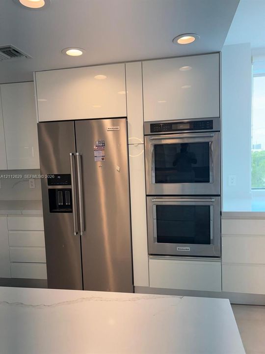 Sleek, high-gloss white cabinetry featuring built-in stainless steel double wall ovens, a matching stainless steel side-by-side refrigerator, recessed ceiling lighting, and solid-surface countertops