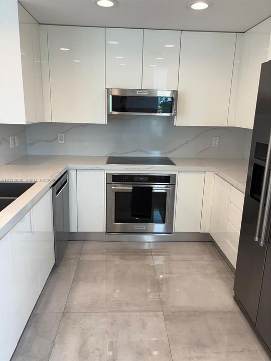 Kitchen featuring high-gloss cabinetry, stone-finish countertops, full-height decorative backsplash, stainless steel appliances, and large-format tile flooring