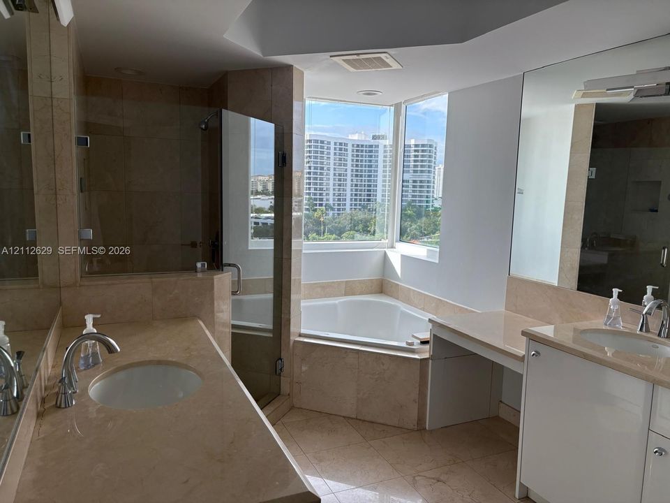 Bathroom featuring dual vanities with stone-topped surfaces, a glass-enclosed shower, a corner soaking tub, and large-format tile flooring
