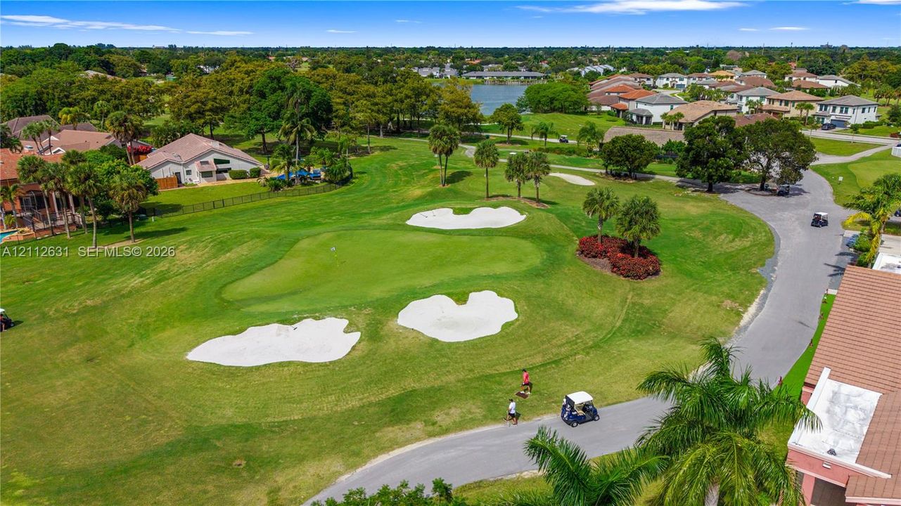 Aerial perspective of a golf course featuring manicured putting greens, sand bunkers, mature palm trees, and water frontage