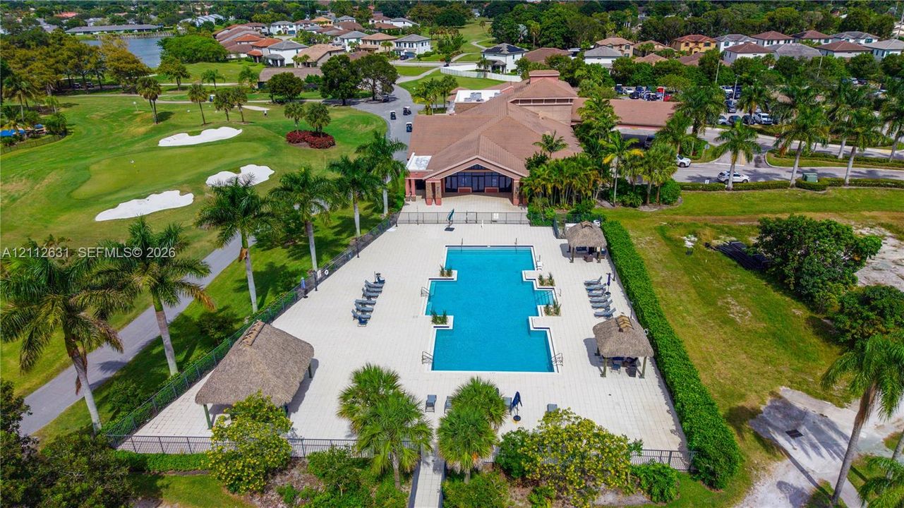 Community pool with paver decking and multiple thatched-roof structures, positioned alongside a golf course featuring manicured greens and sand bunkers
