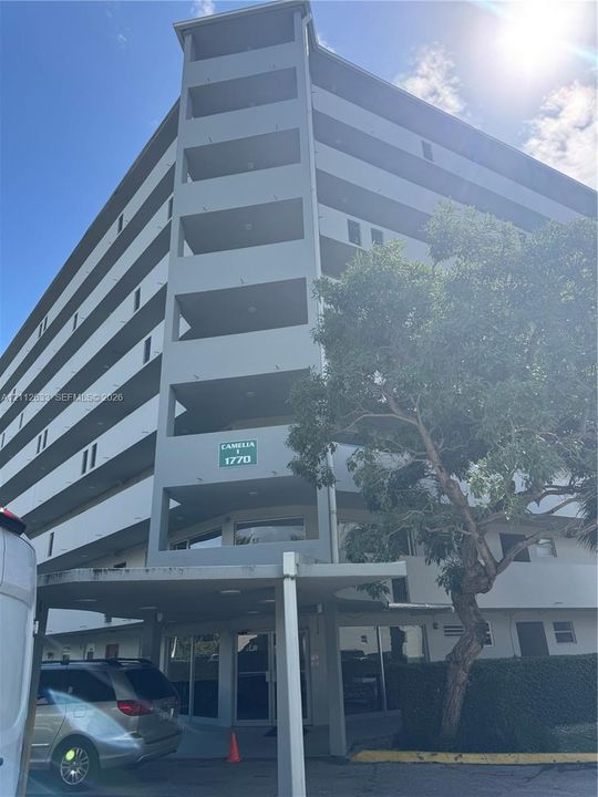 Multi-story residential building featuring covered exterior balconies, a ground-level covered entryway, and mature landscaping