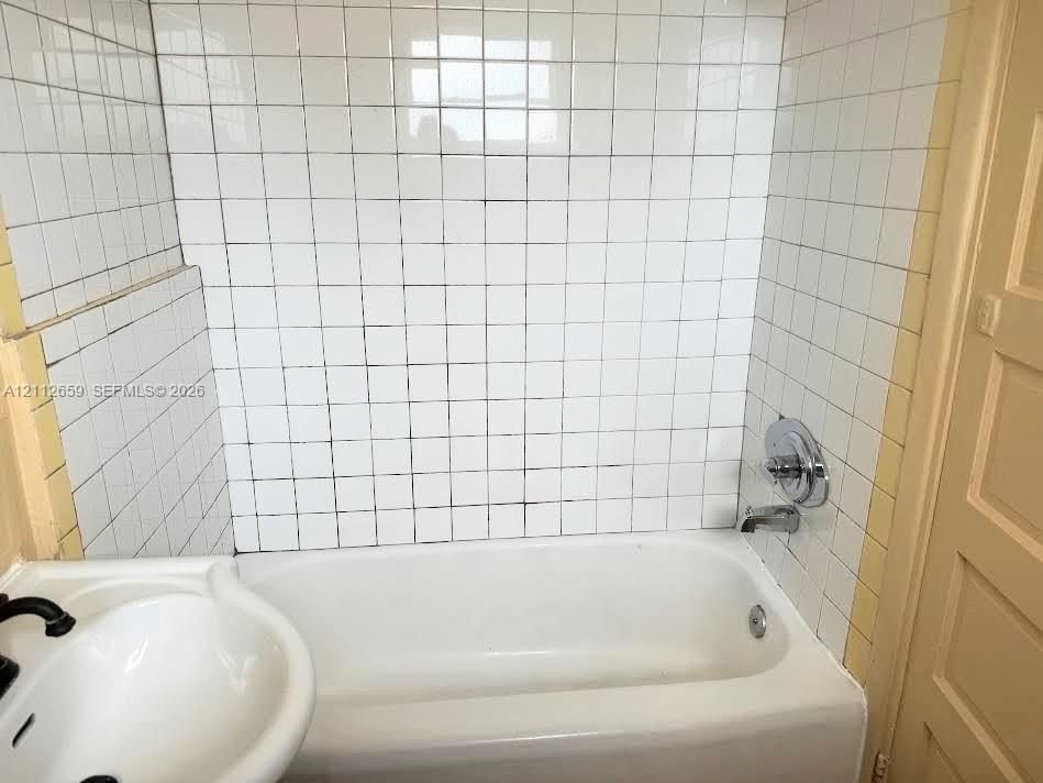 Bathroom featuring a white porcelain tub, square wall tiling, chrome faucet and shower controls, and a pedestal sink with black fixtures