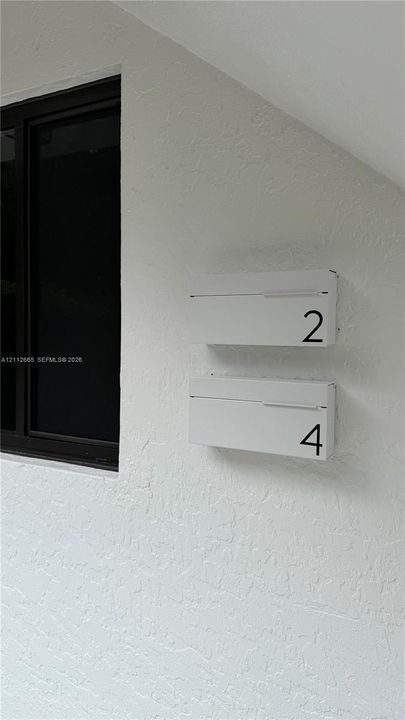 Textured white exterior wall featuring dual minimalist white mailboxes with numerical identification and a black-framed window