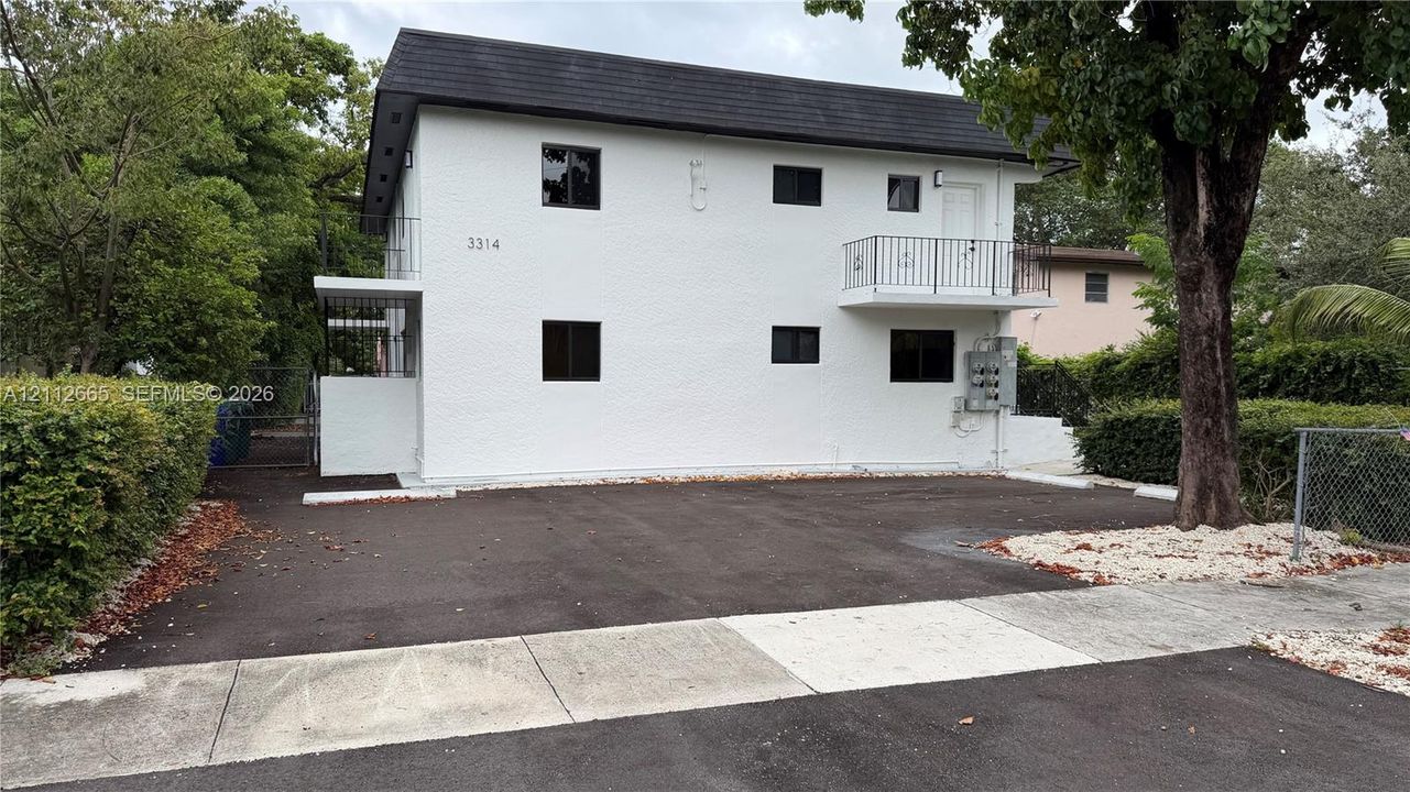 Two-story exterior featuring white stucco finish, asphalt roof, private upper-level balcony with metal railing, and dedicated asphalt parking area west side