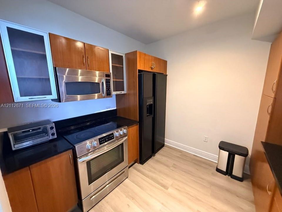 Kitchen featuring wood-finish cabinetry with glass-front accents, stainless steel appliances, dark stone-finish countertops, and wood-finish flooring