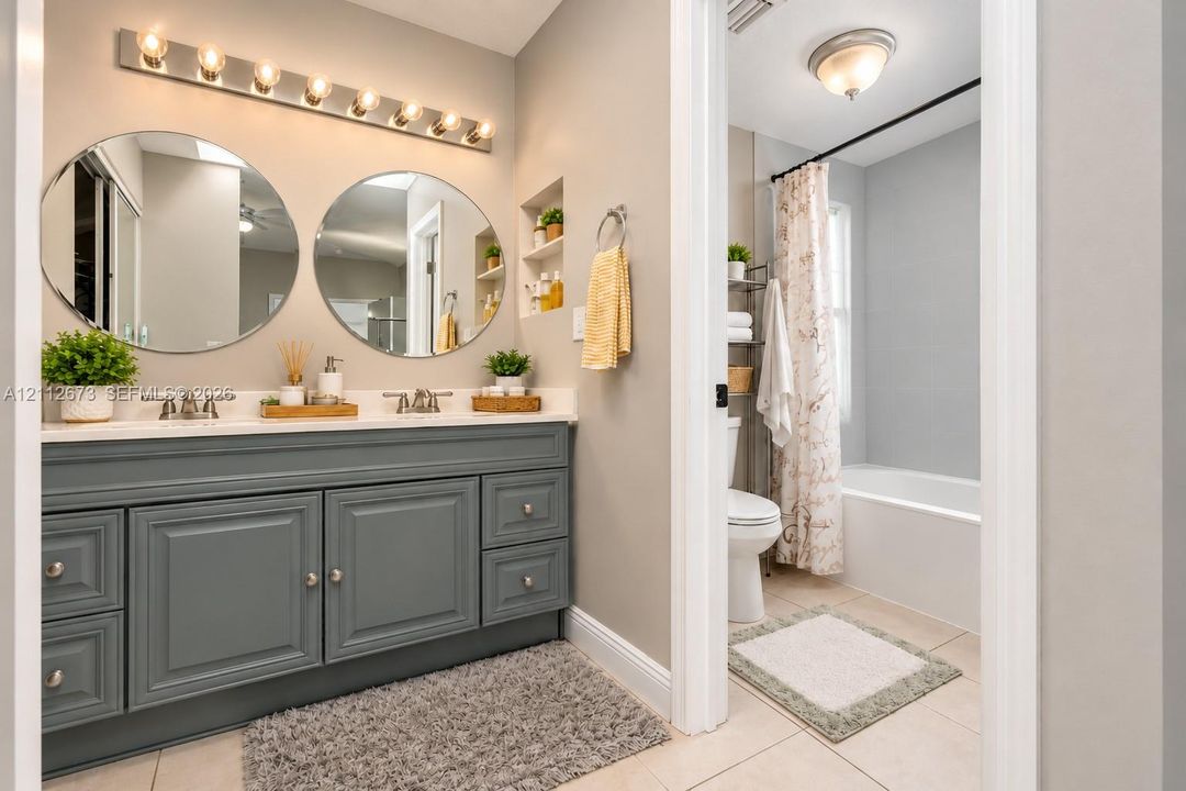 Dual-sink vanity with gray cabinetry and solid-surface countertop, circular wall mirrors, multi-bulb vanity lighting, and recessed wall shelving
