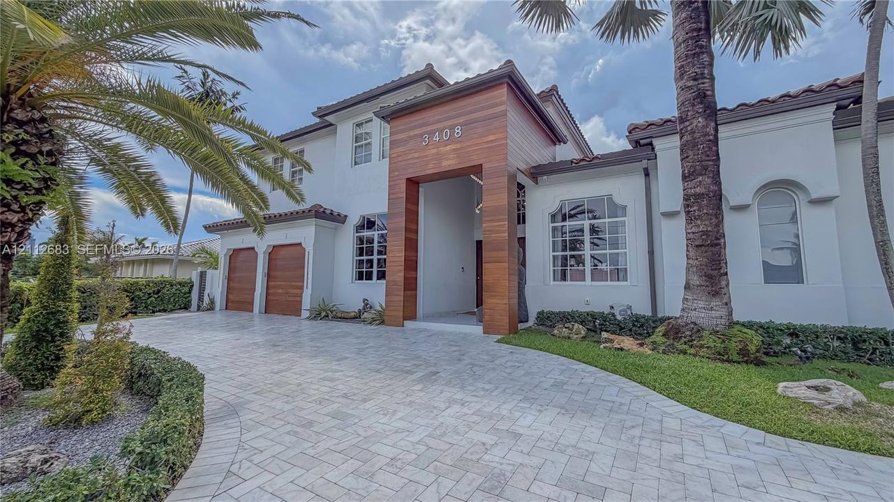 Exterior facade featuring white stucco walls, natural wood-paneled entryway, arched window accents, wood-finish garage doors, and a herringbone-patterned stone paver driveway