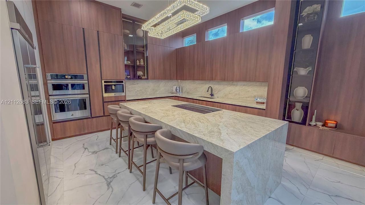 Full-height wood-finish cabinetry with integrated appliances, expansive stone-finish island, chevron-pattern tile flooring, clerestory windows, and architectural linear pendant lighting