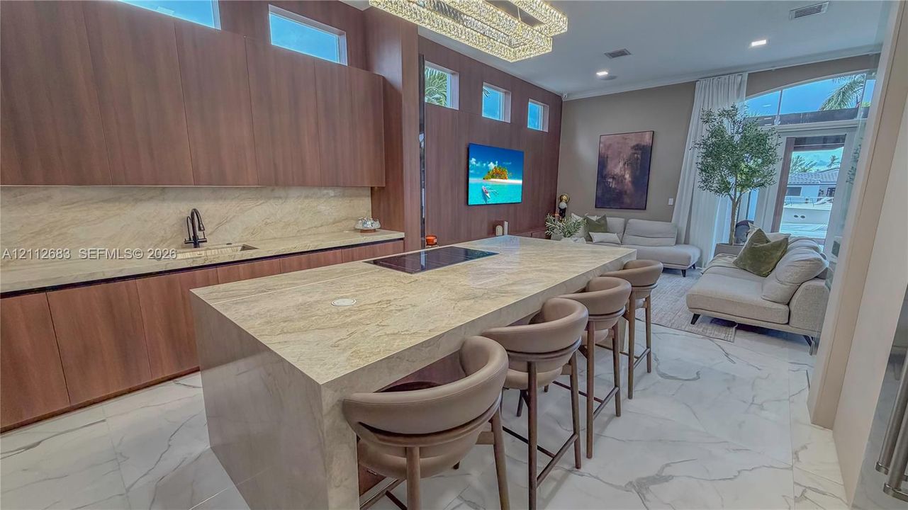 Modern kitchen featuring flat-panel wood-finish cabinetry, a large stone-topped island with an integrated cooktop, stone backsplash, large-format tile flooring, and a linear crystal chandelier