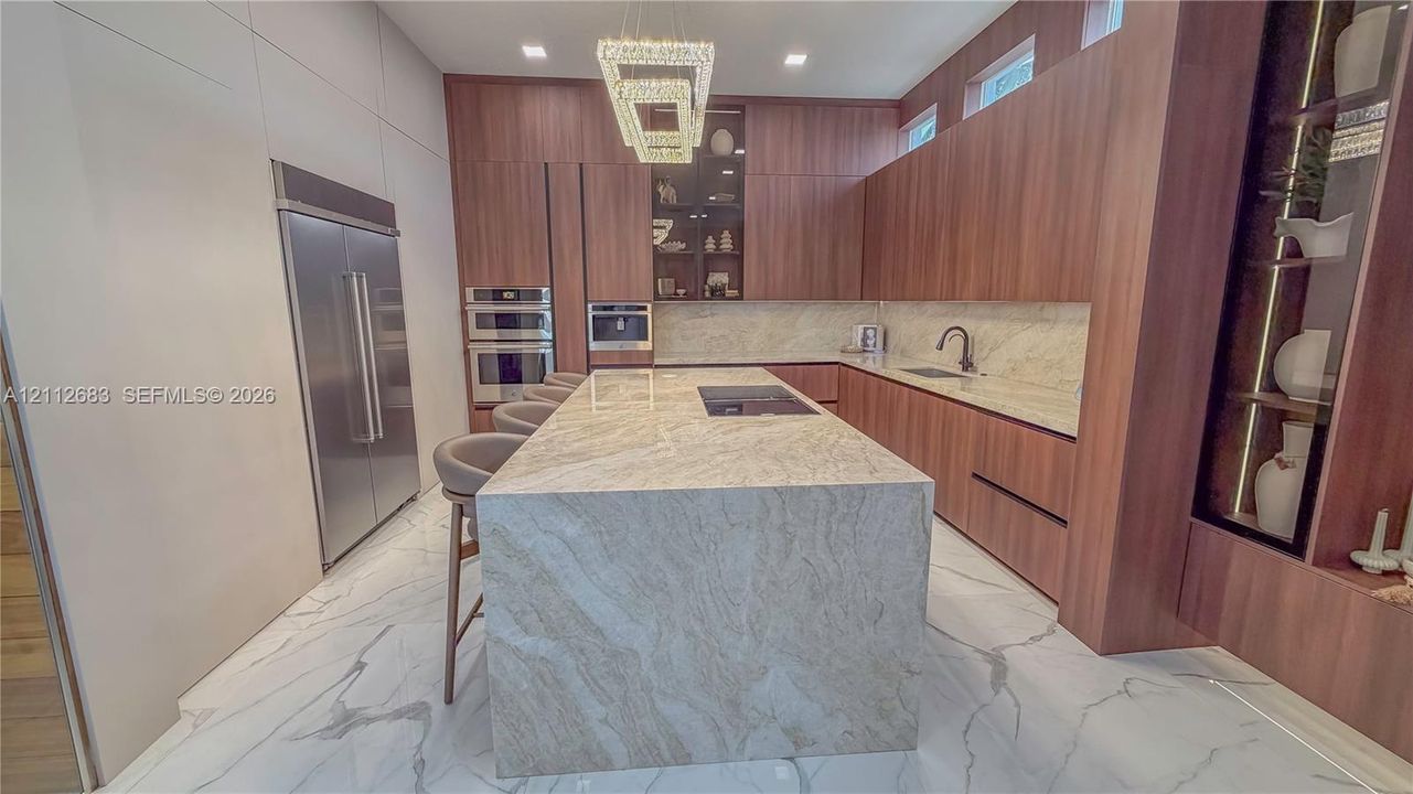 Kitchen featuring wood-finish cabinetry, a waterfall-edge stone island with an integrated cooktop, stone backsplash, built-in stainless steel appliances, and large-format tile flooring