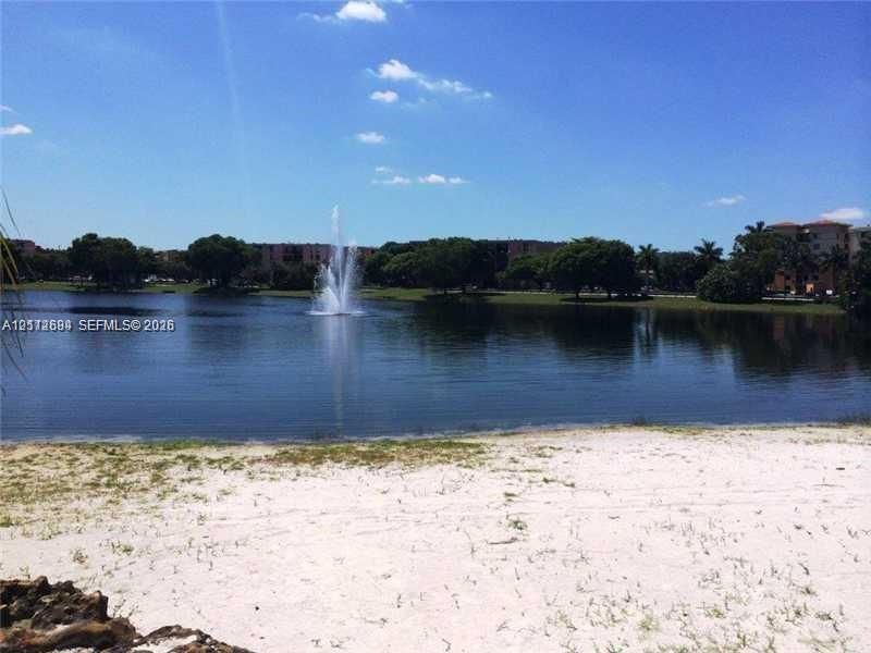 Waterfront setting featuring a central fountain, sandy shoreline, and mature perimeter trees