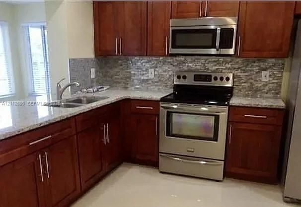 Kitchen featuring rich wood cabinetry, stone-surface countertops, textured tile backsplash, stainless steel appliances, and a dual-basin sink with a pull-down faucet