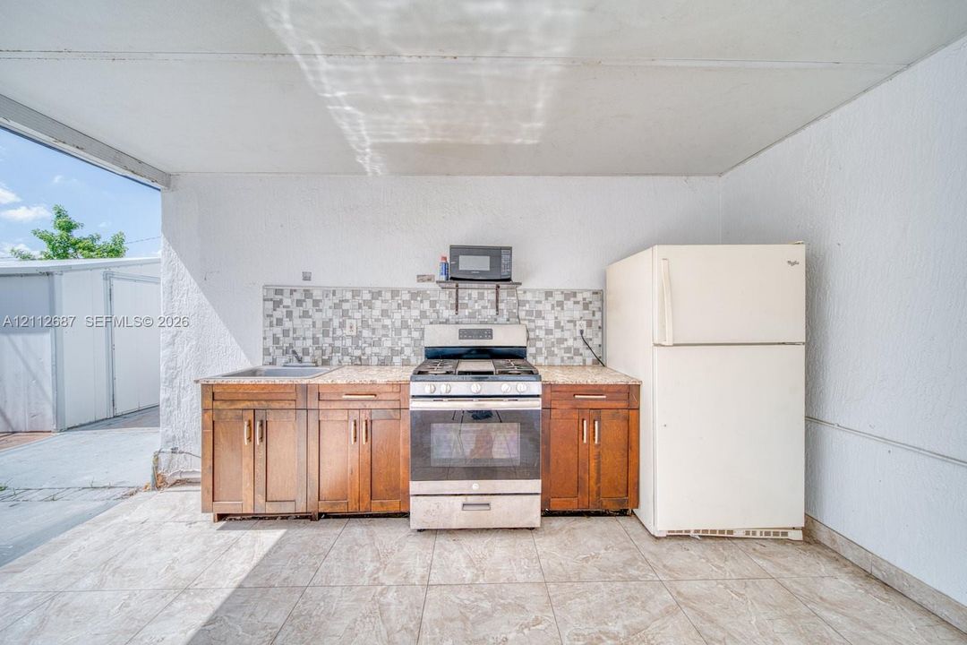 Outdoor kitchen featuring wood-finish cabinetry, stone-look countertop, tiled backsplash, stainless steel gas range, and refrigerator