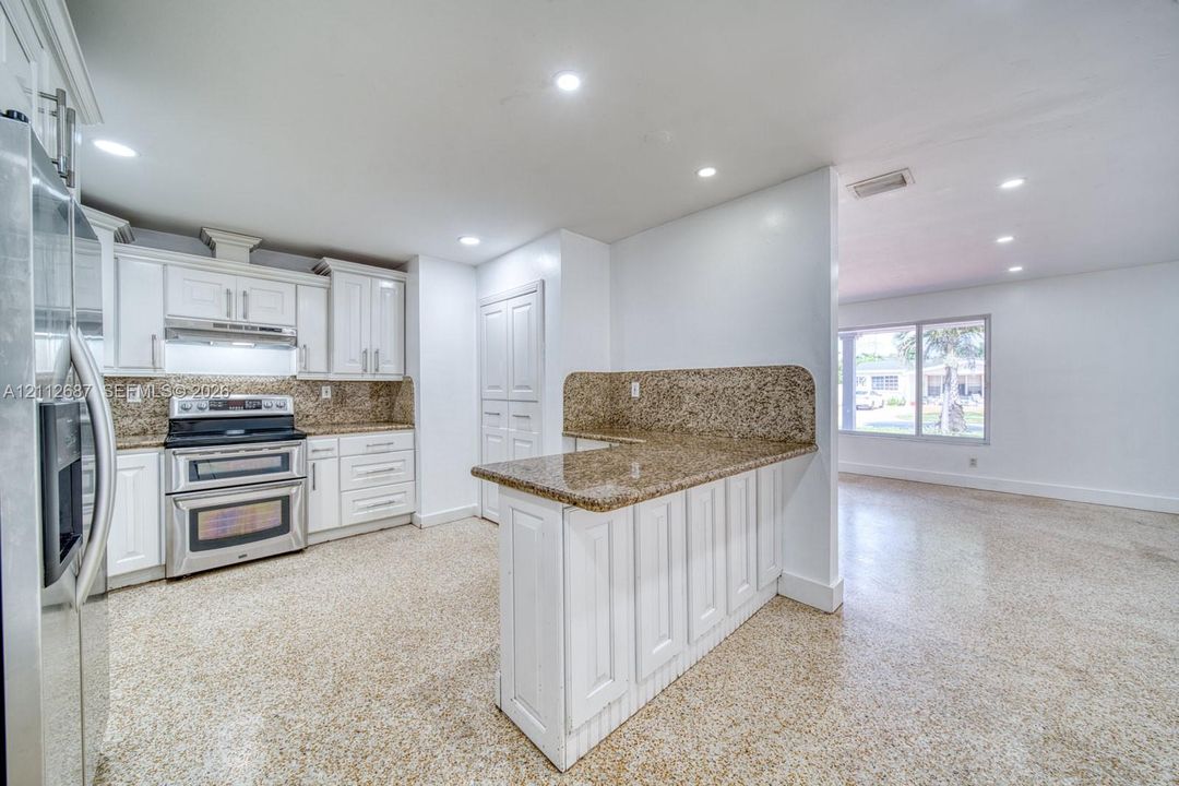 Kitchen featuring white shaker-style cabinetry, stone-surface countertops, stainless steel appliances, a peninsula with bar seating, and polished aggregate flooring