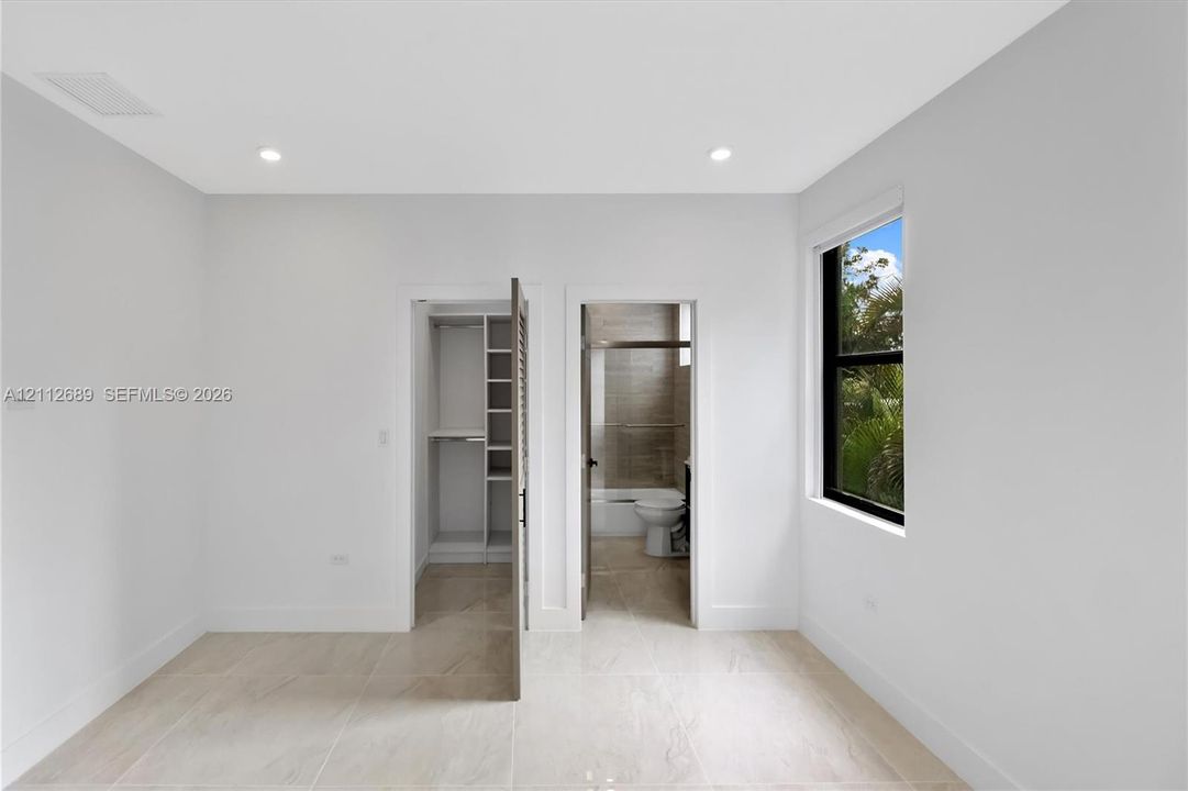Neutral-toned tile flooring, recessed ceiling lighting, a window with a black frame, and a built-in closet featuring shelving and hanging rods
