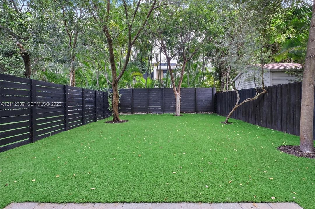 Private backyard featuring horizontal slat fencing, synthetic turf, mature trees, and stone paver border